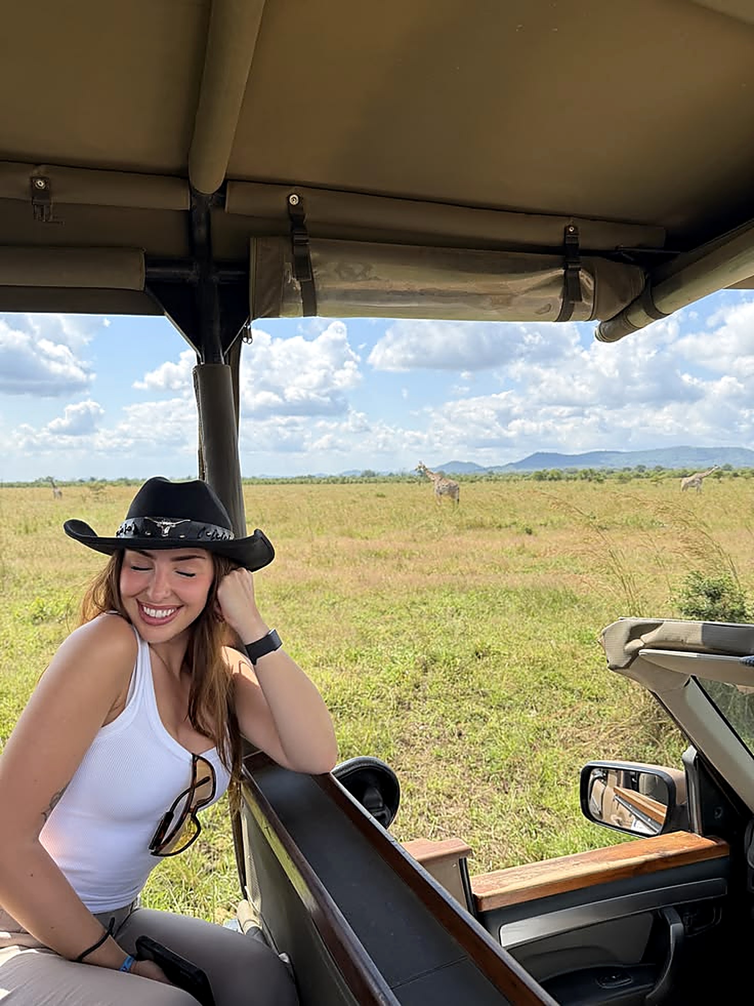 A woman sitting alone on the hood of a safari vehicle, looking out over open grassland under a clear sky